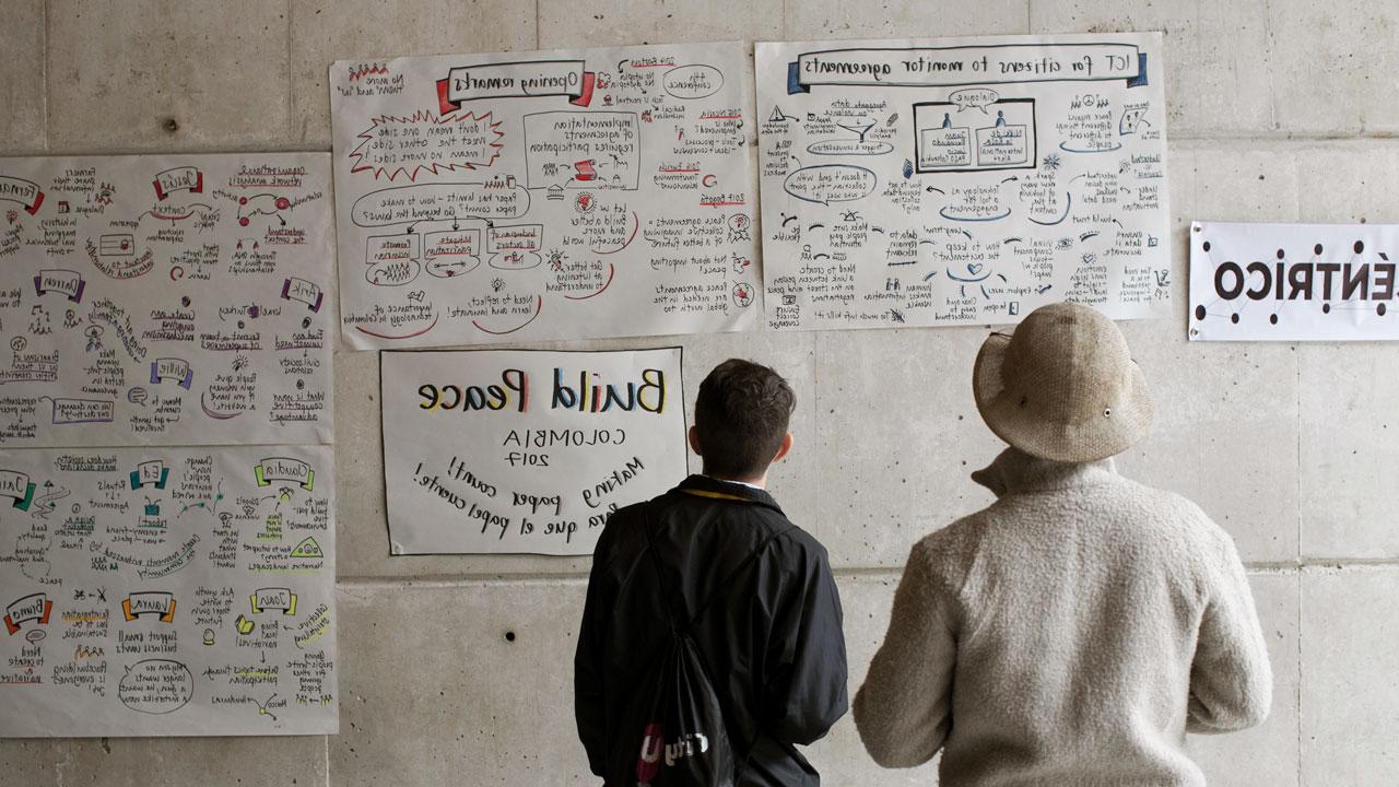 Two people stand in front of a wall with handwritten posters and graphics including one that reads “Build Peace Colombia” as part of Tetra Tech’s community peacebuilding activities