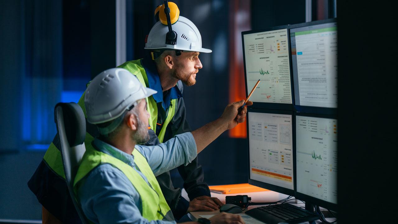 Two people in PPE in an industrial setting look at a bank of computer monitors